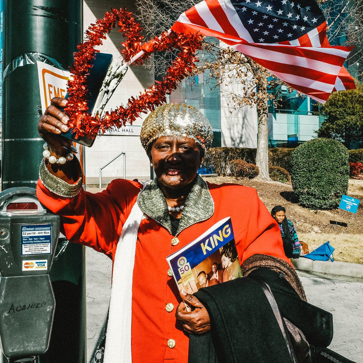 A woman in a red coat holding an American flag and a magazine on a city sidewalk.